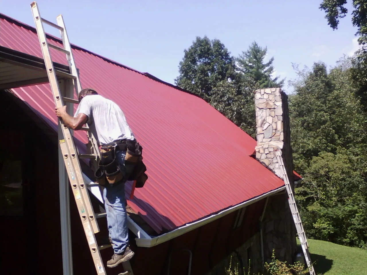 Roofer on a ladder beside a red metal roof and stone chimney — Soffit & Fascia Repair by Roofing Brothers in Williamsville